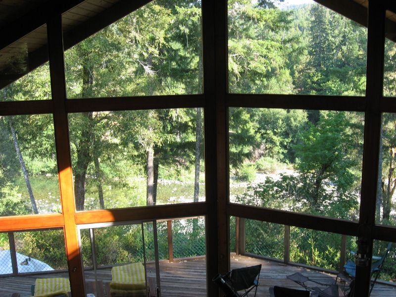 Expansive glass panels provide a view of lush greenery and a flowing river below. Sunlight filters through the trees, casting warm light on the wooden deck. Several outdoor seating options, including yellow-striped lounge chairs, are visible, inviting relaxation in the natural surroundings.