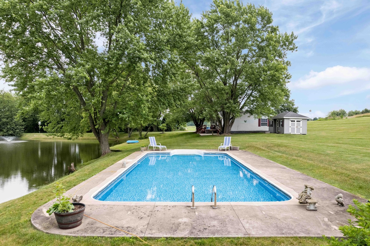 A serene swimming pool is surrounded by grassy areas and mature trees, reflecting the blue sky. Two lounge chairs are positioned beside the pool, and the adjacent pond is visible in the background, creating a peaceful outdoor atmosphere.