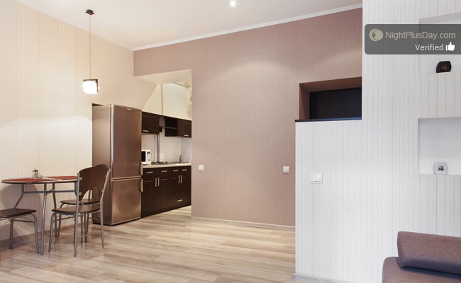 A functional kitchen is visible, featuring sleek cabinetry and modern appliances. A small dining table with two chairs is positioned nearby. The living area showcases light wooden flooring and pale wall finishes, creating a bright and airy feel throughout the space.