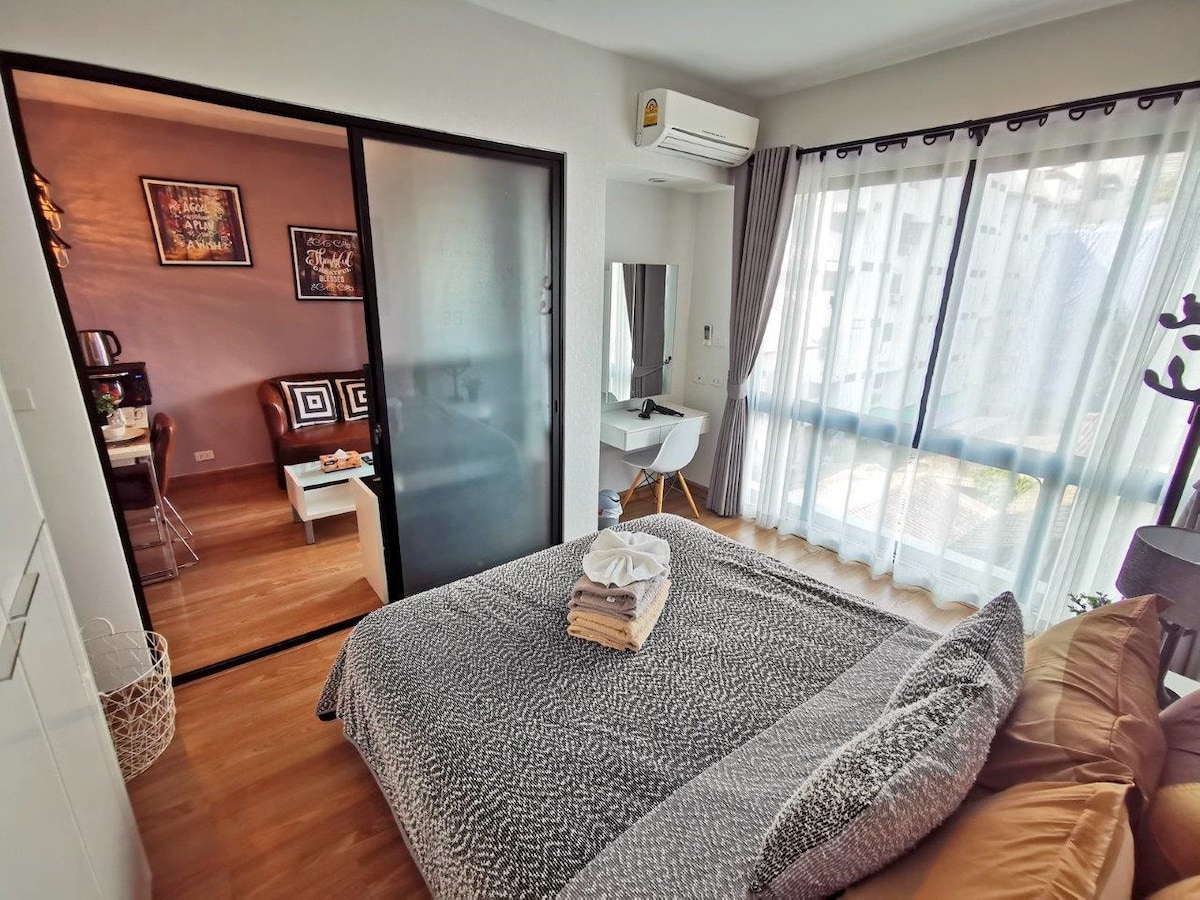 A modern bedroom is highlighted by a spacious bed with neatly arranged linens. Natural light filters through large glass windows, showcasing a comfortable workspace and a stylish chair. A sliding door adds privacy while maintaining a contemporary aesthetic in the welcoming space.