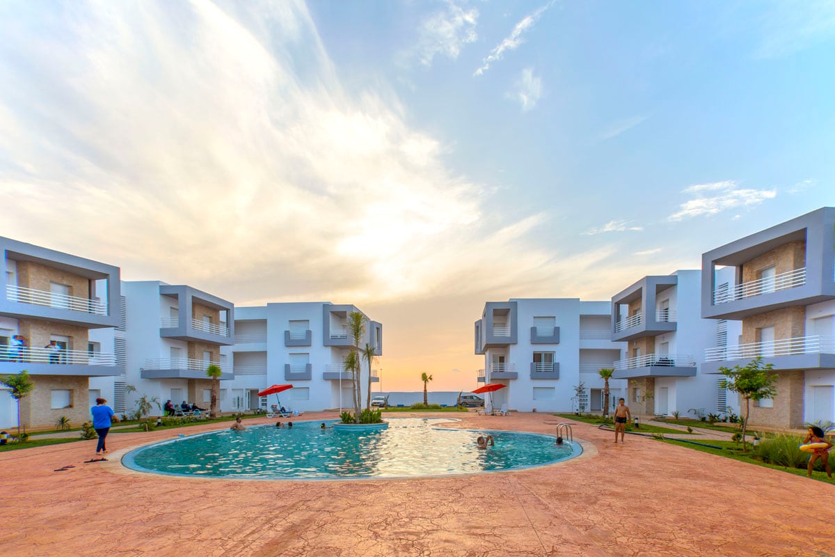 A serene view of the residence showcases a central pool surrounded by white buildings. Guests can be seen enjoying the water. Comfortable lounge chairs and umbrellas are positioned around the pool area, reflecting a welcoming and communal atmosphere.