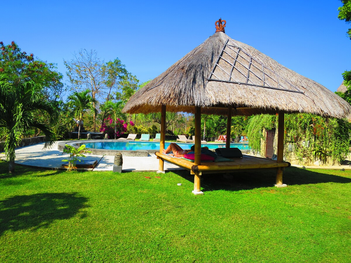 A thatched roof gazebo is set in a lush green garden, providing shade next to a large swimming pool. Palm trees and colorful foliage surround the area, with sun loungers positioned by the water, inviting relaxation in a serene outdoor setting.