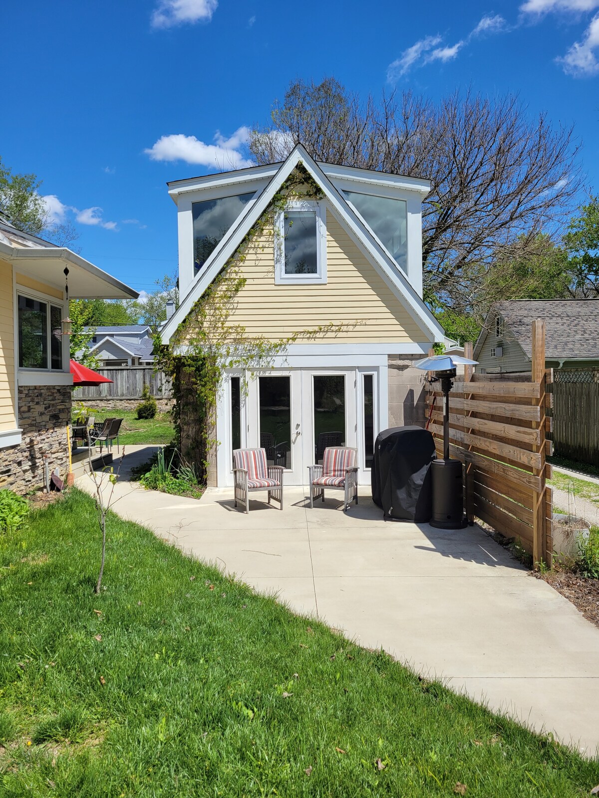 The exterior of the granny flat is visible, featuring a charming A-frame design with large windows. Two striped chairs are positioned on a concrete patio, surrounded by lush greenery and a wooden privacy screen. Bright blue skies and fluffy clouds create a cheerful atmosphere.