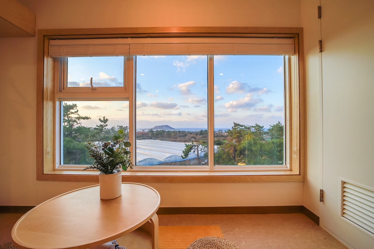 A large window provides a panoramic view of the landscape, featuring distant mountains and a blue sky dotted with clouds. A small round table with a potted plant stands in the foreground, inviting natural light into the space.
