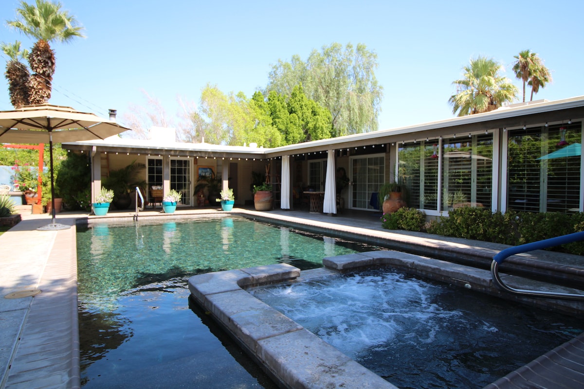 A serene outdoor area features a clear swimming pool complemented by a heated spa. Surrounding greenery and potted plants provide a natural ambiance. The area is bordered by a shaded patio with umbrellas, ensuring a peaceful space for relaxation.