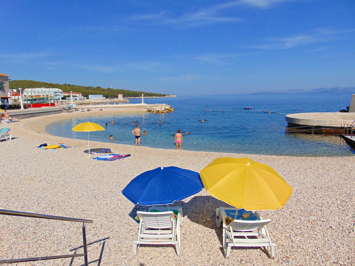 Two lounge chairs are positioned under a blue and a yellow umbrella on a pebbled beach. Clear blue waters stretch towards the horizon, where swimmers can be seen enjoying the sun. Nearby, a promenade features beachgoers and a rocky shoreline.