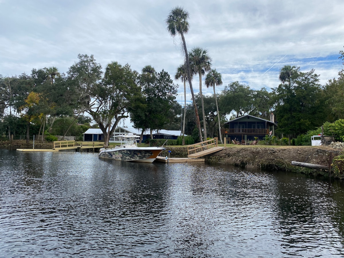 The riverfront view showcases a spacious dock with a boat moored nearby. Lush palm trees line the shore, and the two-story lodge is located in the background. The water reflects the surrounding greenery, creating a serene atmosphere.