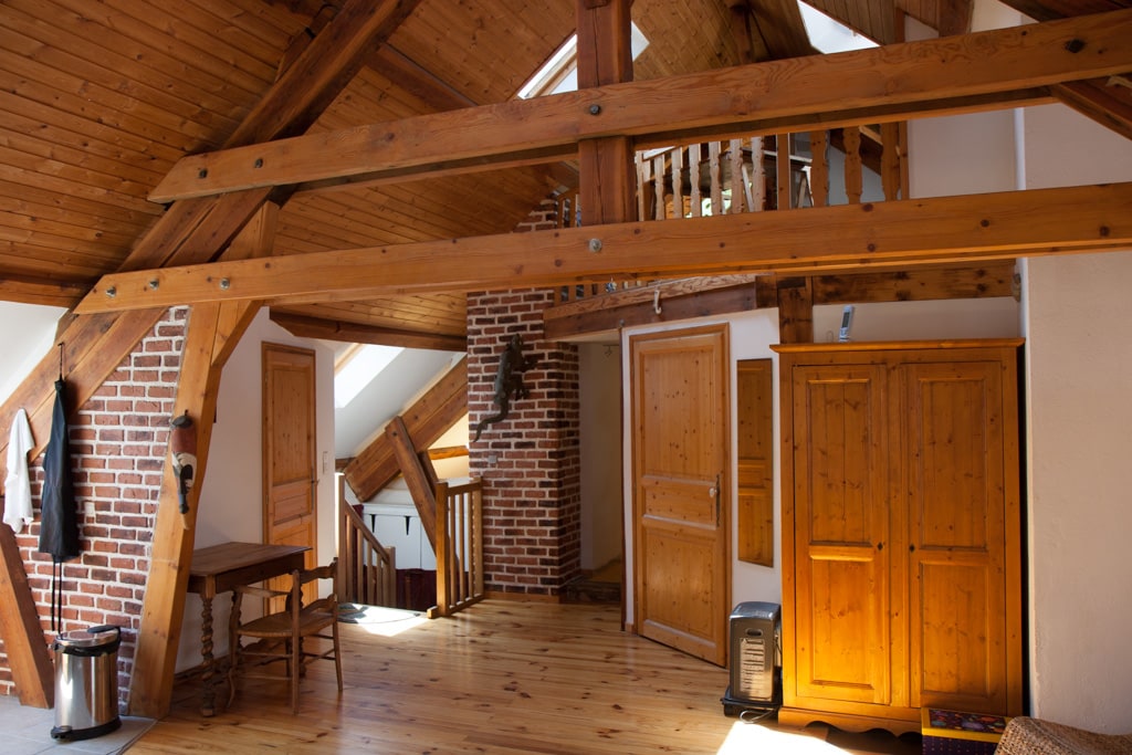 The open living space features exposed wooden beams and a combination of wooden and brick finishes, enhancing the character of the duplex. A wooden dining table and chairs are positioned by a window, while a wardrobe stands against the wall. Natural light fills the area from multiple skylights.
