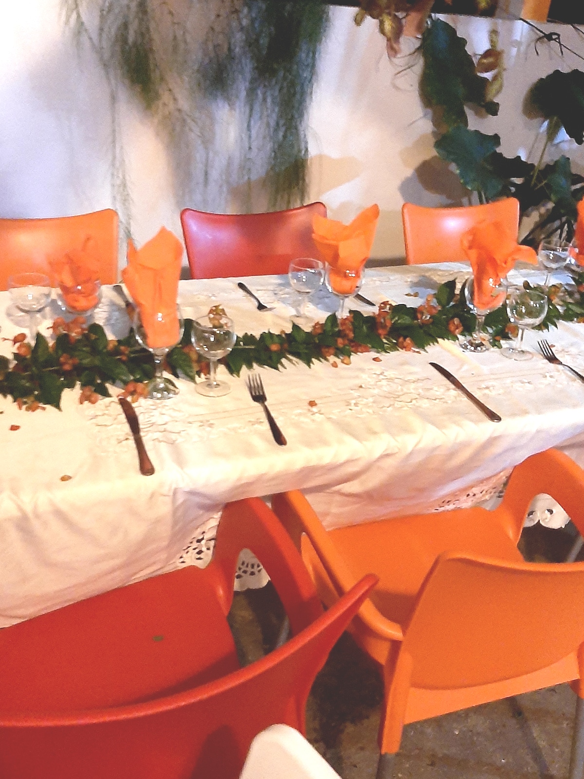 A dining table is set with a white tablecloth, adorned with greenery and scattered petals. Orange napkins are artfully arranged at each place setting. Surrounding chairs in vibrant shades of orange complement the setup, creating a lively and inviting atmosphere.