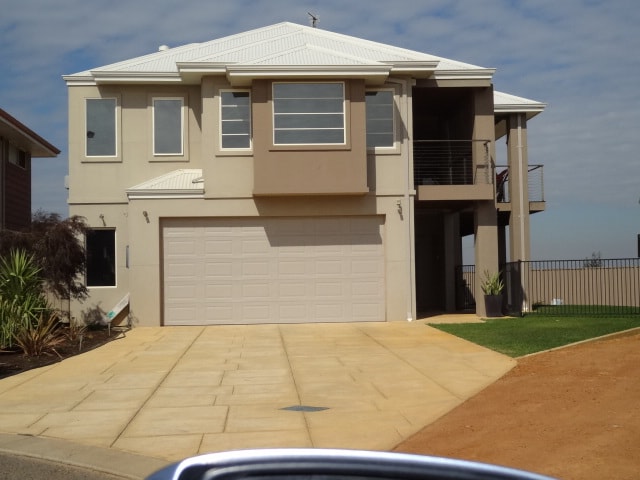 A modern two-story house is shown with a light-colored exterior, featuring large windows and a balcony. The garage occupies part of the ground floor, and the driveway is spacious, leading to a well-maintained lawn. The surrounding area is residential, with additional homes in view.