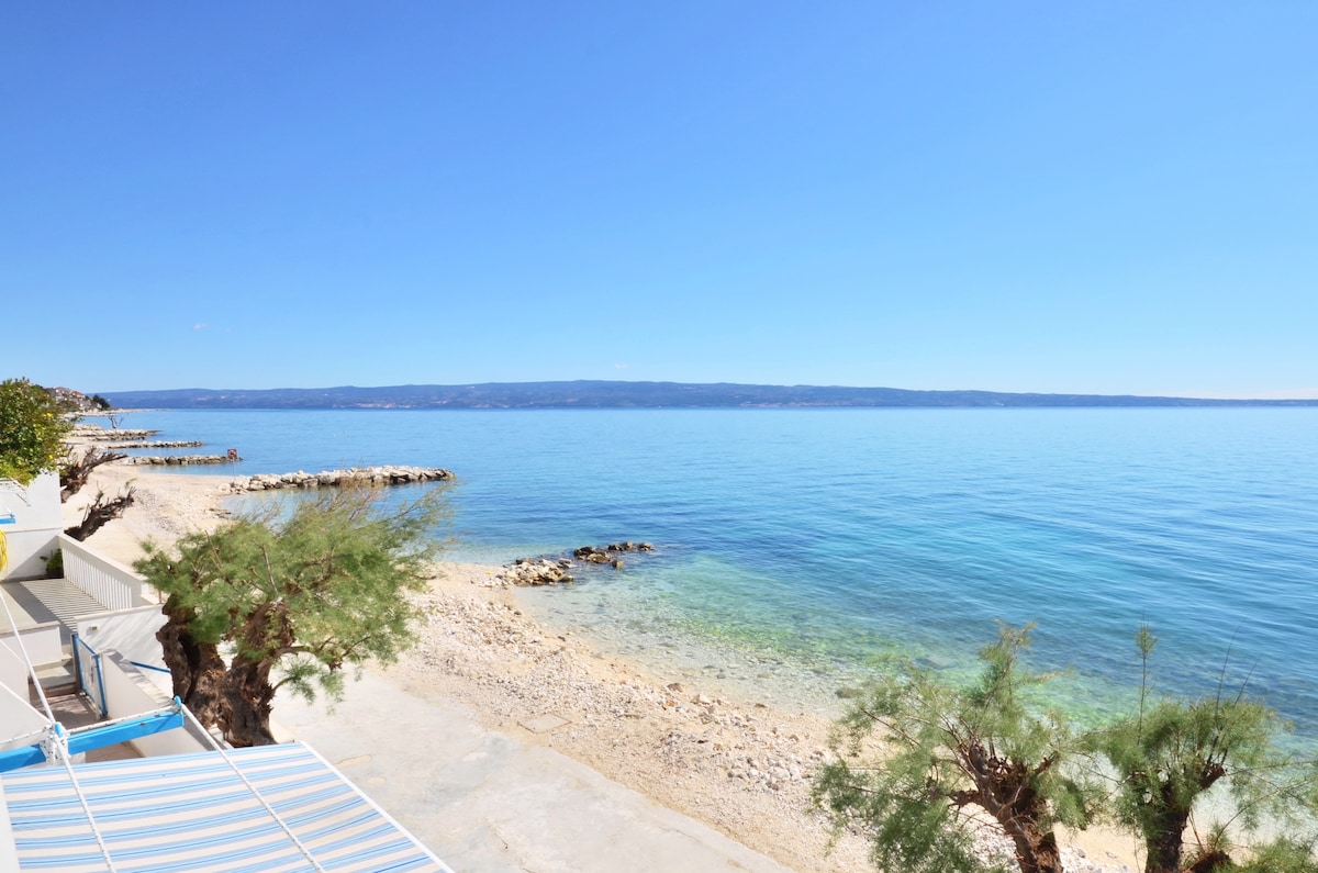 A serene view of a pebble beach is presented, with calm turquoise waters extending toward distant hills. The clear sky complements the tranquil scene, creating an inviting atmosphere for relaxation by the sea. Lush vegetation lining the shore adds a touch of greenery.