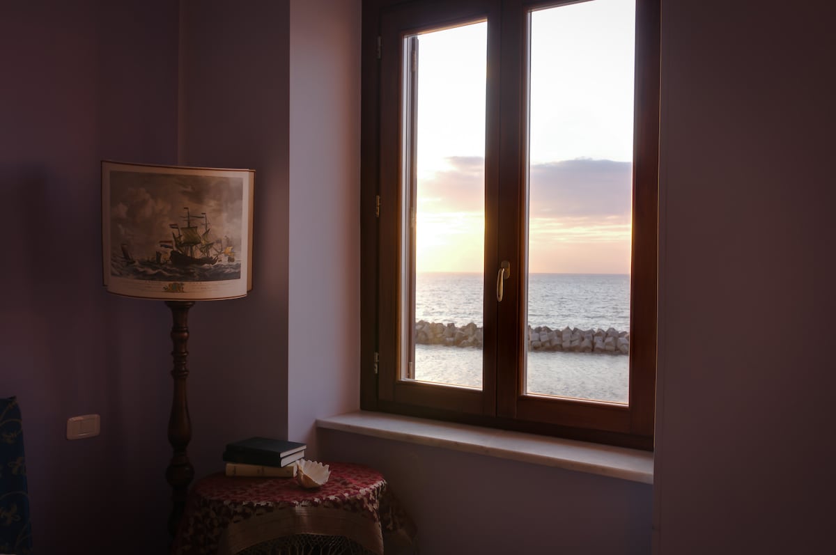 A window frames a view of the sea at sunset, with shades of orange and purple reflected on the water. A lamp and a stack of books decorate the nearby table, creating an inviting ambiance in this elegantly finished room.