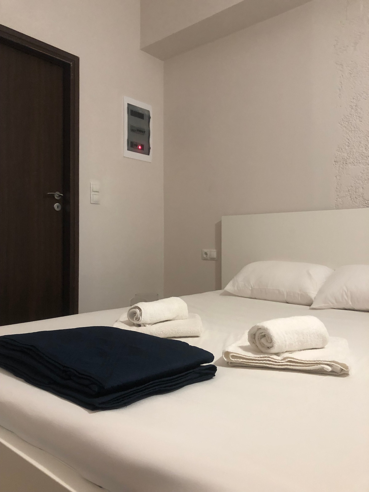 A cozy sleeping area is presented, featuring a spacious bed with white linens and neatly arranged towels in varying sizes. A dark blue blanket is folded at the foot of the bed, and a wooden door is visible, adding to the room's inviting atmosphere.