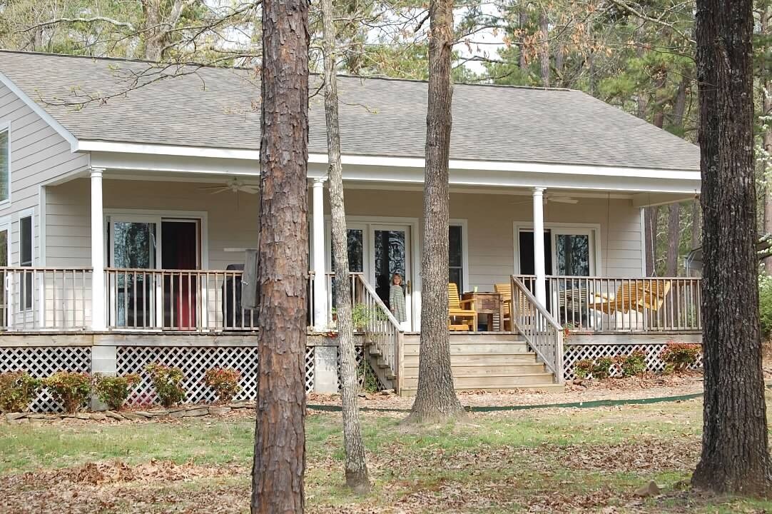 The lake house features a welcoming covered and uncovered deck area surrounded by trees. Several seating options are visible, with a few chairs placed on the deck. The exterior displays a light-colored facade with sliding doors leading to the interior.