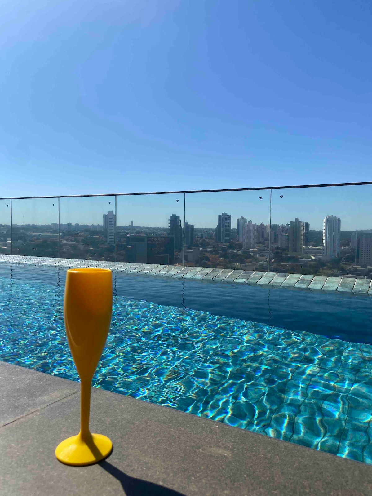 A vibrant yellow glass is positioned at the edge of an infinity pool, reflecting sunlight. The clear blue water of the pool creates a serene scene, while a panoramic view of the city skyline extends in the background under a clear blue sky.