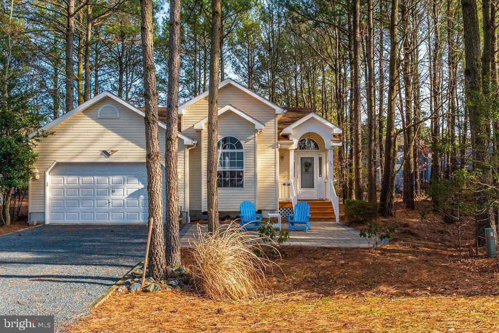 The exterior of a three-bedroom home is showcased amidst tall pine trees. A gravel driveway leads to a garage, while two blue chairs are positioned on the front steps by the entrance, inviting relaxation in the natural surroundings.