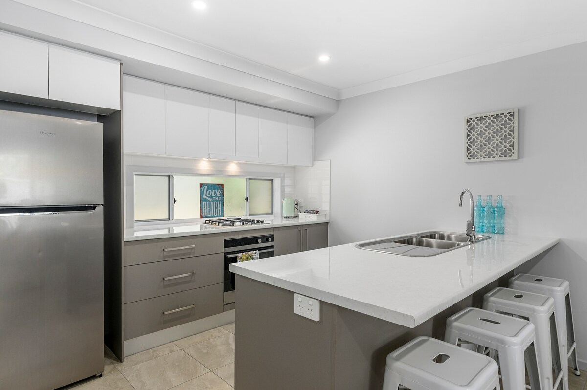 The modern kitchen features sleek cabinetry and a spacious countertop with bar seating for four. Stainless steel appliances are visible, including a refrigerator and cooking facilities. A window allows natural light to illuminate the area, enhancing the room's functional layout.