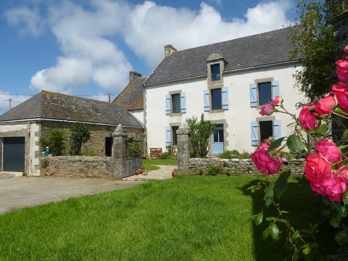 A large farmhouse is set against a bright sky, featuring blue shutters and a neatly trimmed garden. A pathway leads to the entrance, surrounded by greenery. Vibrant flowers bloom in the foreground, enhancing the welcoming appearance of the property.