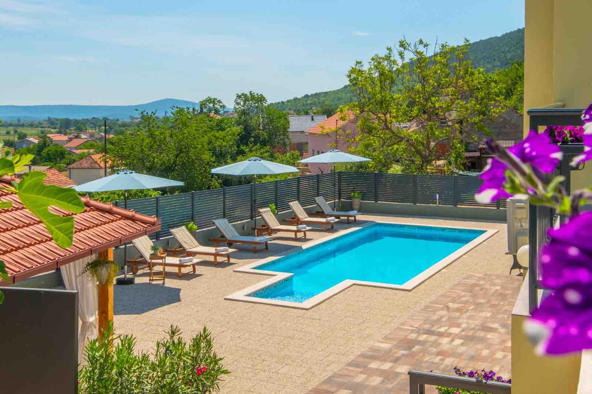 An outdoor area features a sparkling pool surrounded by comfortable loungers with umbrellas. Lush greenery and colorful flowers contribute to a refreshing ambiance, while the view extends to the distant hills and village homes, creating a serene and inviting setting.