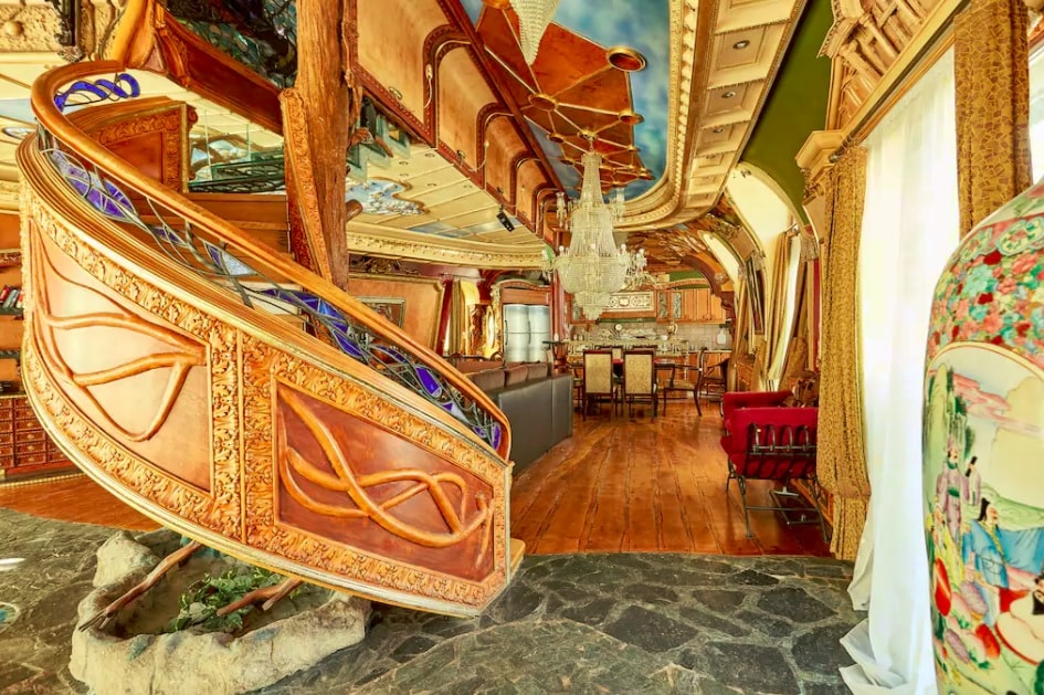 An intricately designed staircase, featuring ornate wooden details, curves gracefully towards the upper level. A chandelier with crystal accents hangs overhead, illuminating the spacious dining area with elegant furnishings. Natural stone flooring complements the rich textures present throughout the expansive living space.