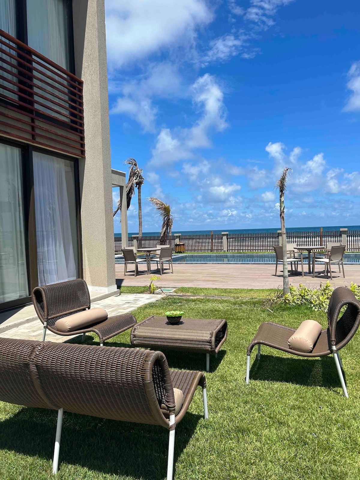 An outdoor seating area is presented with several modern chairs and low tables, set on lush green grass. The backdrop features a view of the sea and a clear blue sky, enhancing the tranquil ambiance of this beachside location.