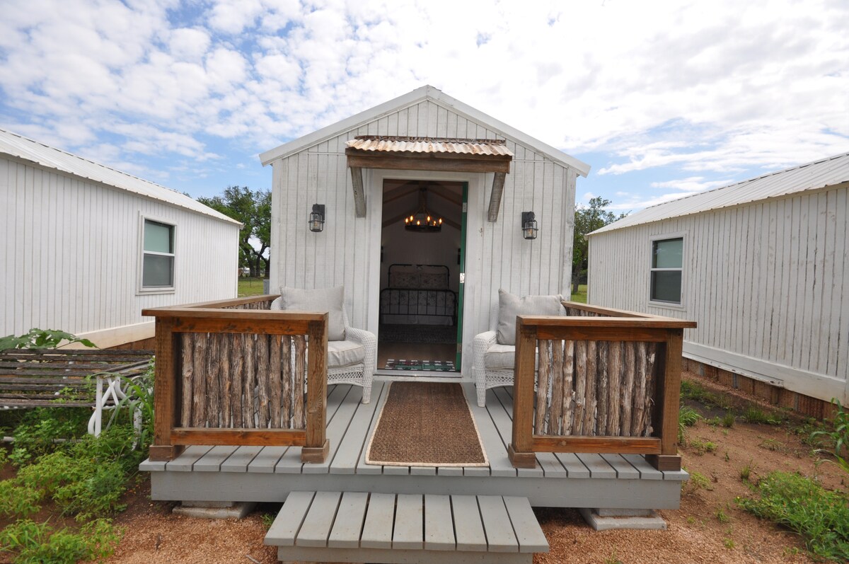 A quaint cabin entrance is framed by two rustic wooden chairs on a small porch. The cabin features a light-colored facade, with large windows on either side. A welcoming doormat lies at the doorstep, set against a backdrop of green grass and a cloudy sky overhead.