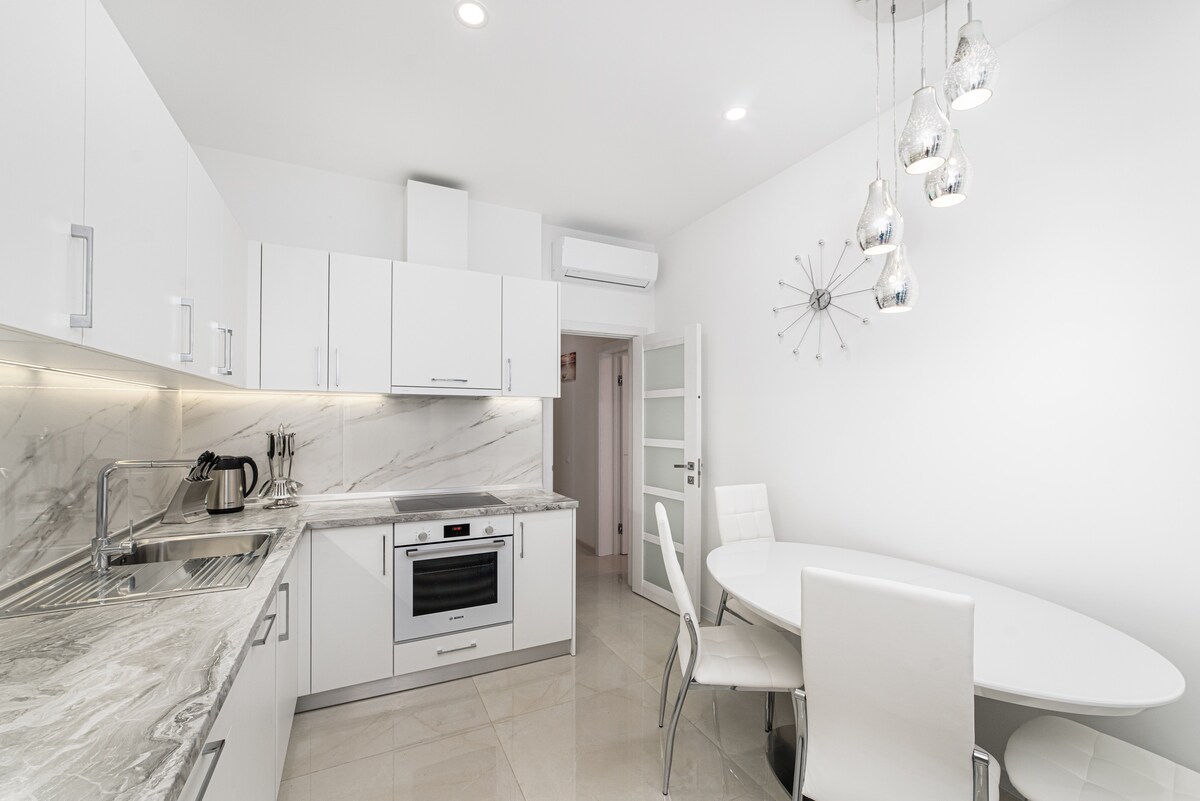 A modern kitchen features sleek white cabinetry and marble countertops. Stainless steel appliances include an oven and a microwave. A round dining table is surrounded by white chairs, with pendant lights hanging above, illuminating the space with soft light.