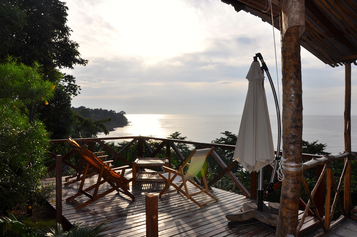 A wooden deck offers a stunning view of the sea, featuring a light-colored umbrella and several chairs arranged for relaxation. Lush greenery surrounds the area, enhancing the natural ambiance, while a soft light illuminates the horizon where the sea meets the sky.