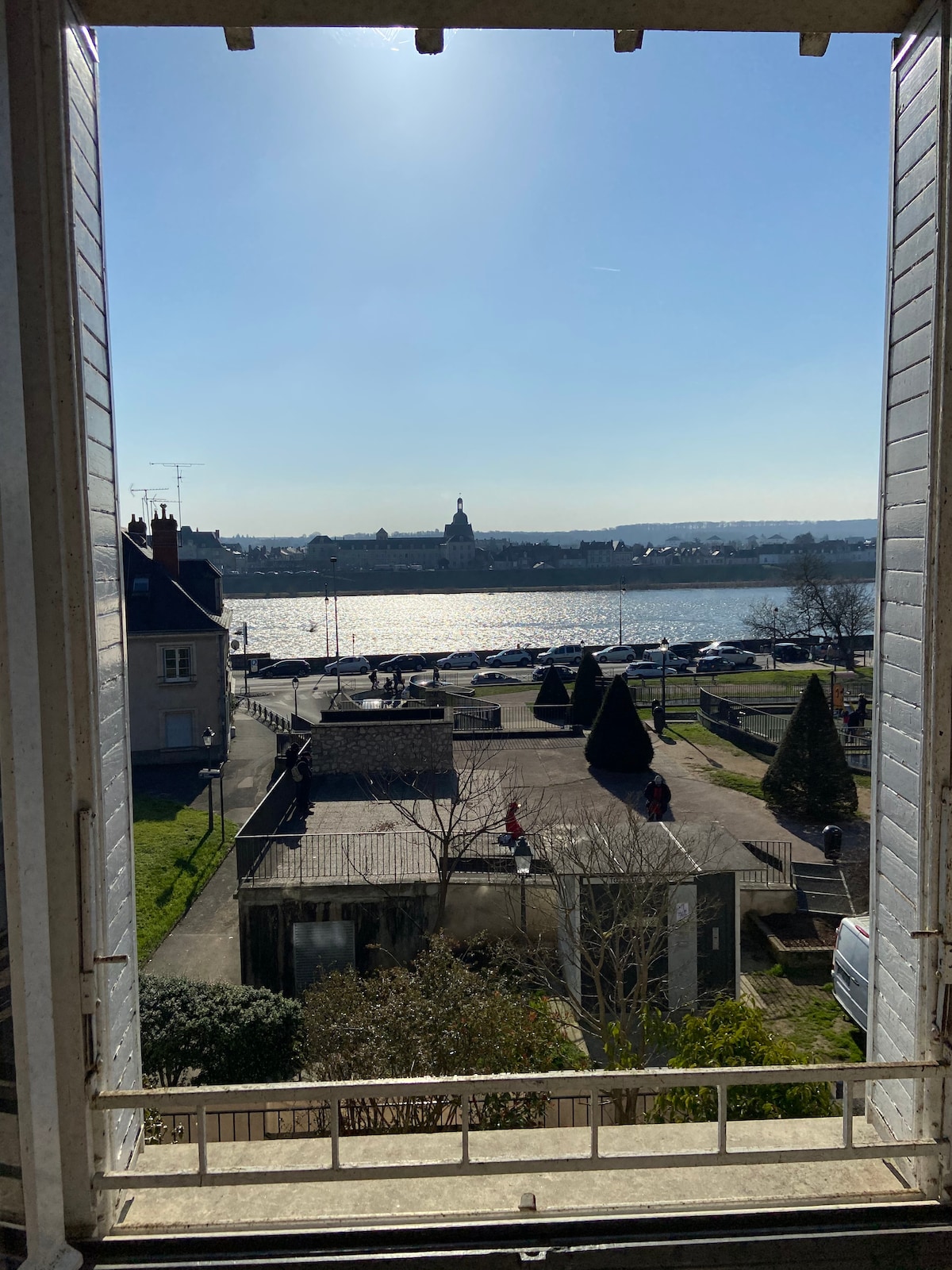 A wide view is framed by an open window, revealing the shimmering Loire River under sunlight. The scene includes a landscaped area with neatly trimmed trees and a pathway, while distant buildings are visible across the river.