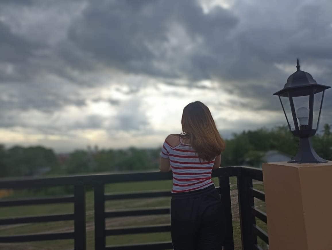 A person stands on a balcony overlooking a cloudy sky, framed by a lantern on the right. The view includes expansive greenery in the background, enhancing the sense of open space and tranquility.