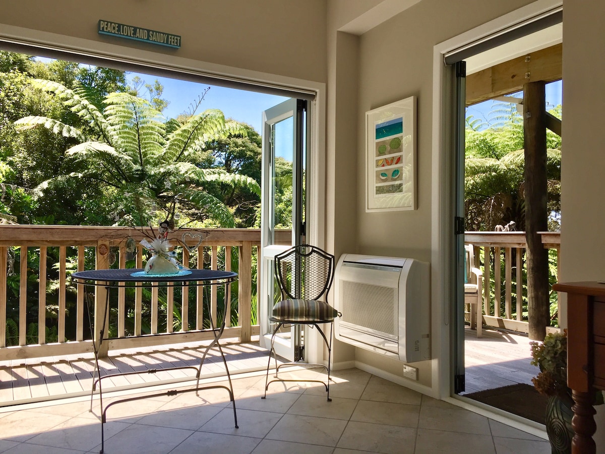A light and airy lounge area is presented, featuring large glass doors that open to a private deck. A small table with chairs is positioned near the entrance, while lush greenery is visible outside, creating a seamless connection to the natural surroundings.