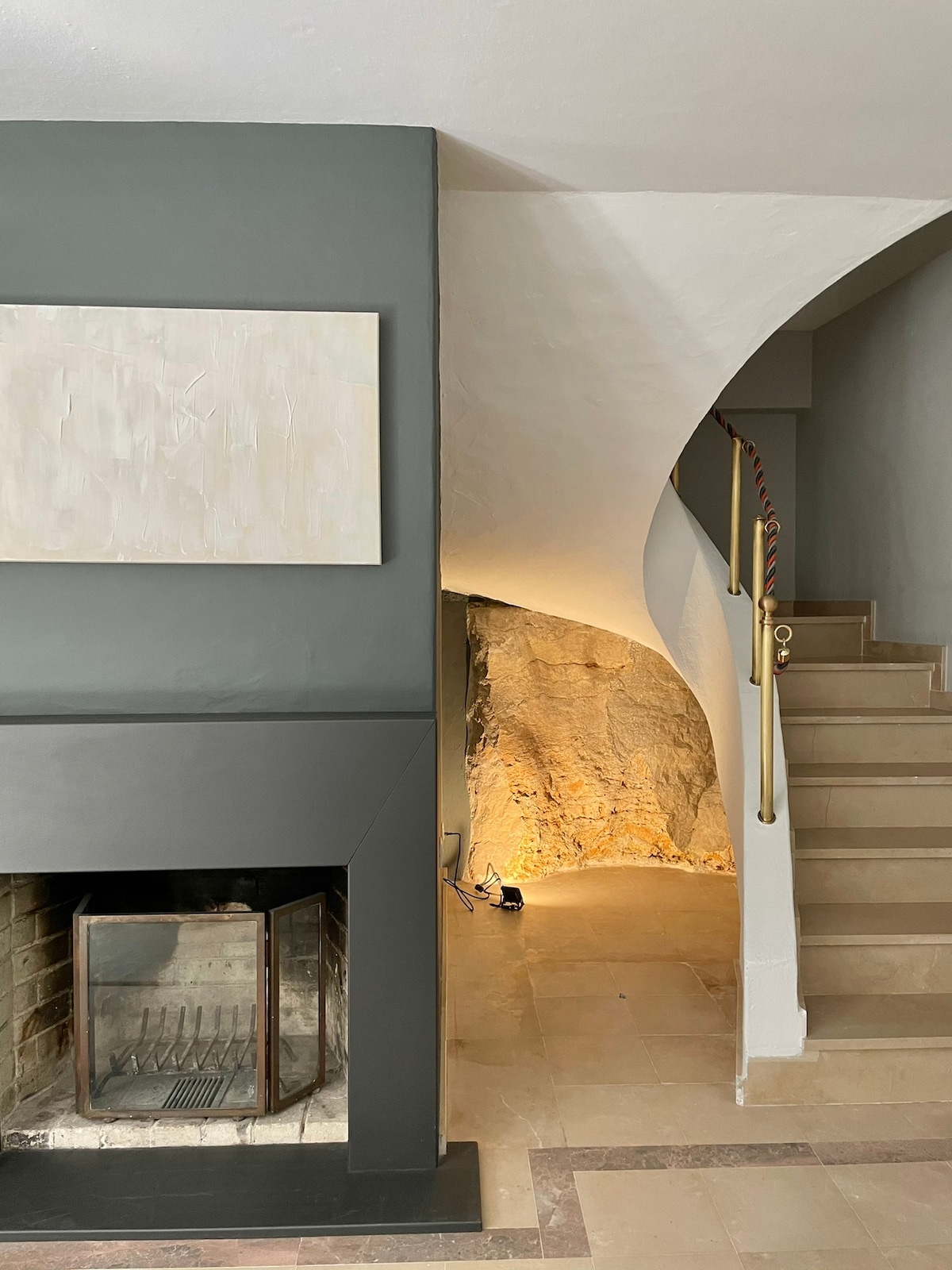 A minimalist interior features a curved staircase leading upward. A modern fireplace is positioned against a grey wall, complemented by a textured stone wall partially illuminated by soft lighting. Warm tones enhance the overall ambiance of the space.