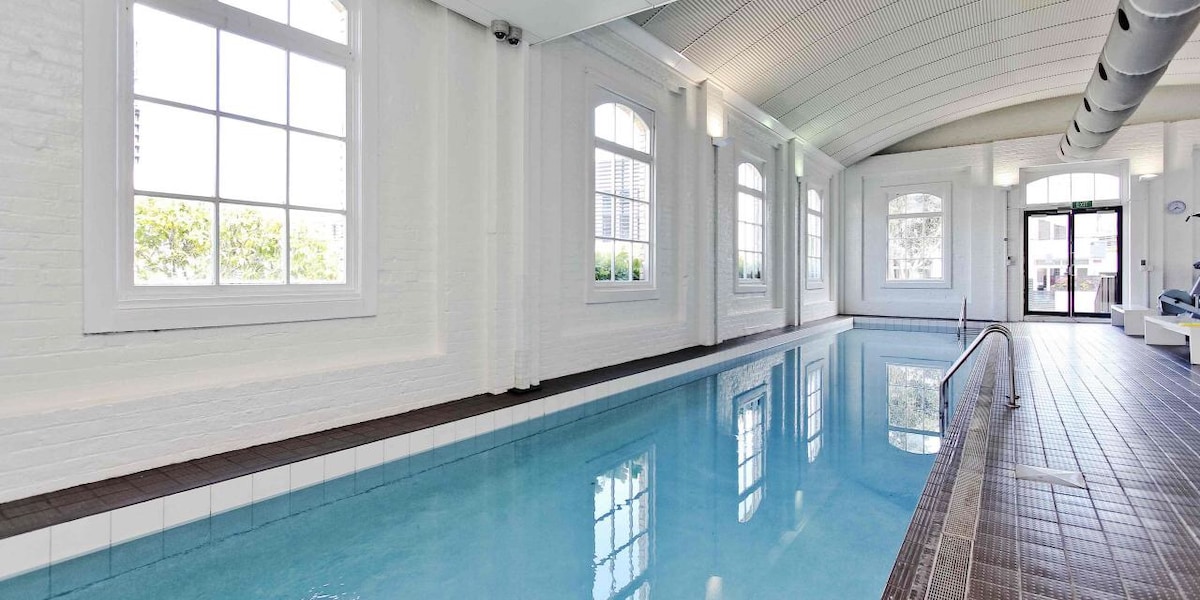 An indoor swimming pool is visible, featuring a clear blue surface reflecting natural light from large windows. White brick walls contribute to a bright atmosphere, while a glass door leads to an outdoor area. The pool area is designed for relaxation and leisure.