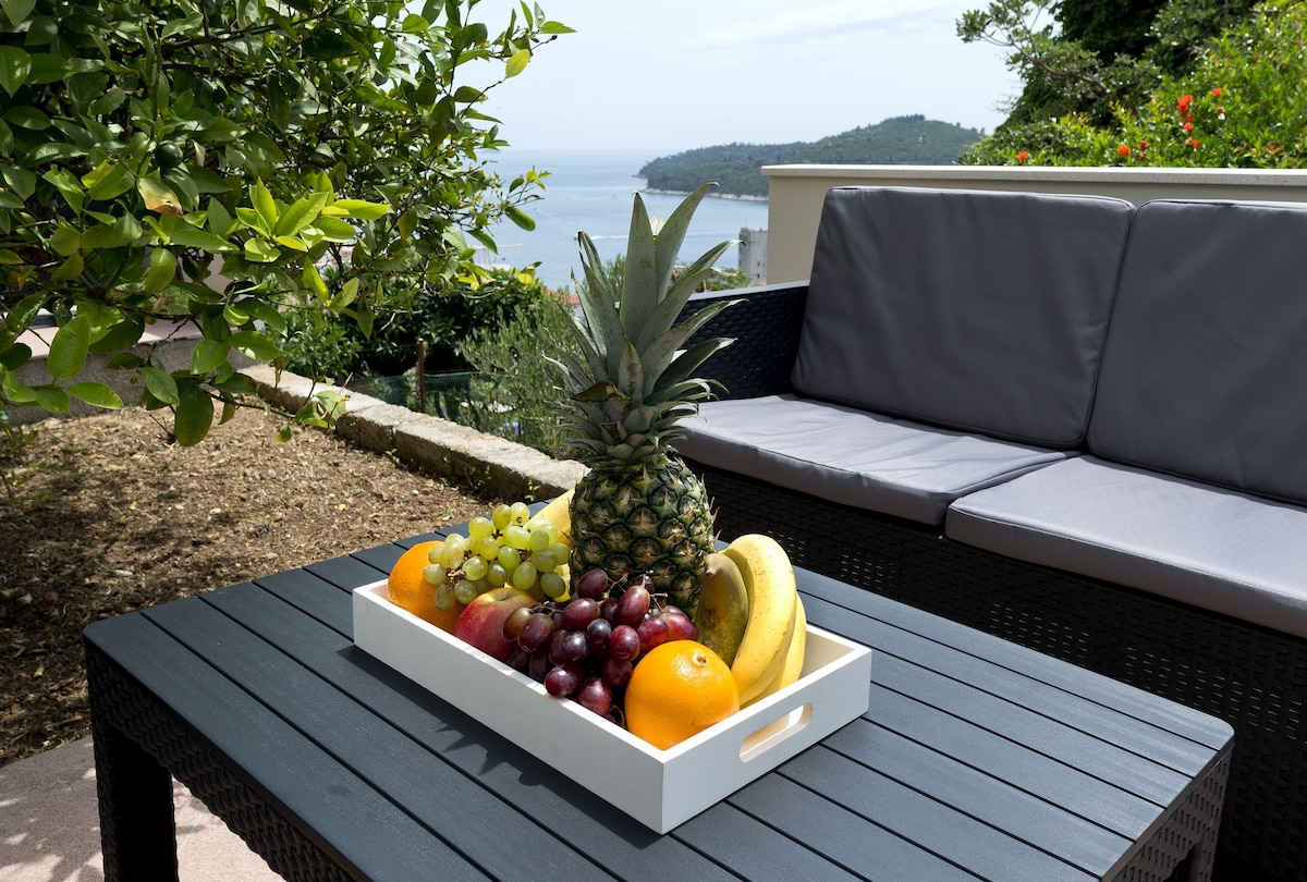 A wicker table is accompanied by a comfortable seating area, featuring a gray sofa. A white tray holds a variety of fresh fruits, including a pineapple, grapes, bananas, an orange, and other seasonal items. In the background, a coastal view is visible.