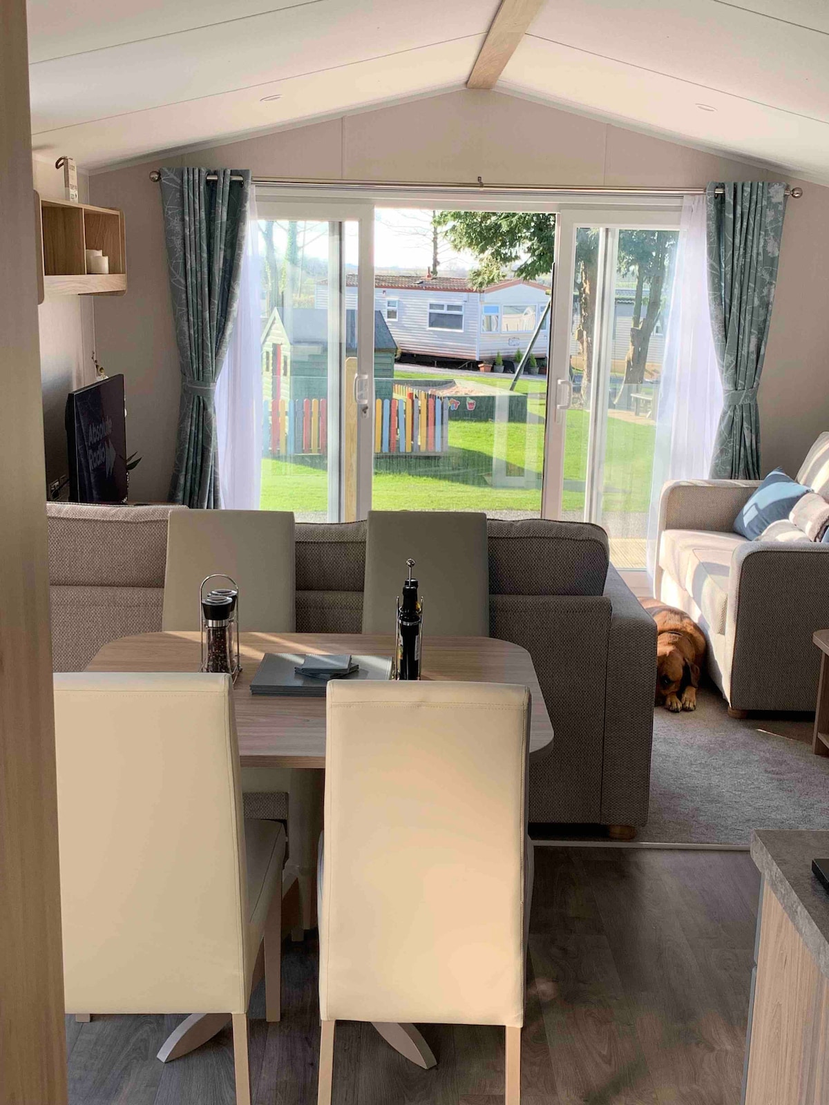 An open-plan layout features a dining table with light-colored chairs, positioned in the foreground. A spacious lounge area with a grey sofa offers views through large glass doors to an outdoor grass area, where colorful play equipment is visible. Natural light fills the room.