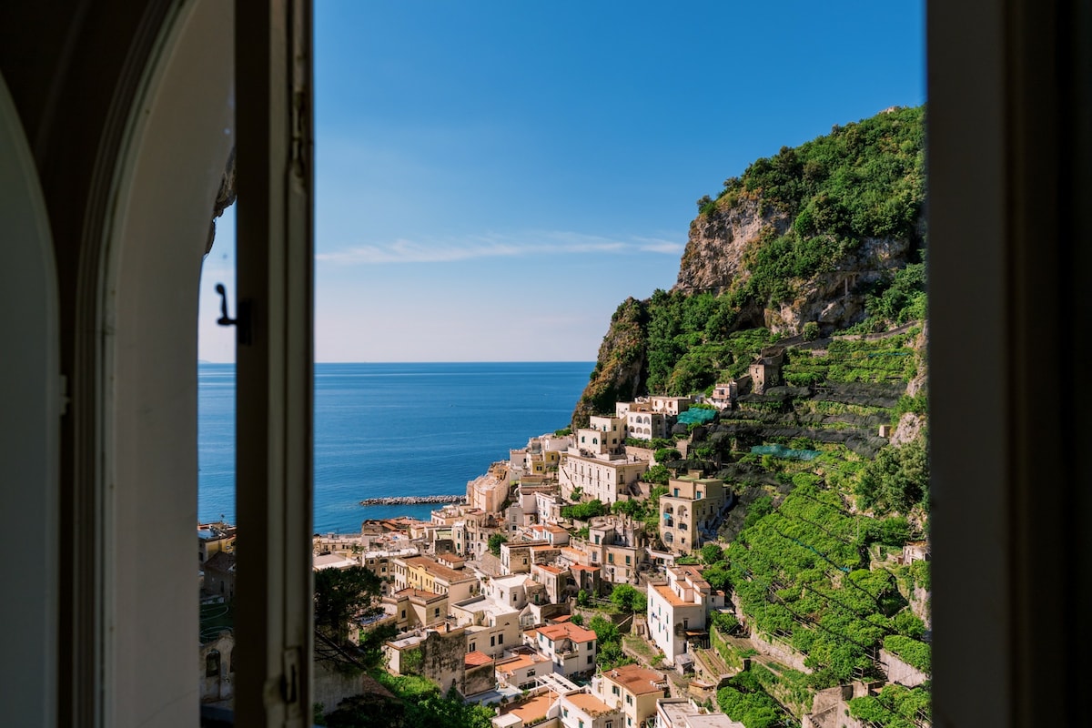 A serene view of the coastal village is framed by an open window, showcasing terraced greenery and traditional Mediterranean architecture. The deep blue sea extends to the horizon under a clear sky, reflecting the charm of the Amalfi Coast.