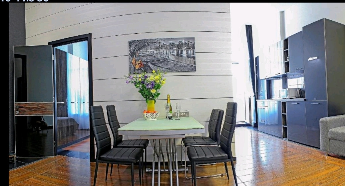 A modern dining area features a rectangular table set with a vase of flowers, surrounded by six black chairs. A painting depicting a cityscape adorns the wall, and a doorway leads to another room. The kitchen area is visible in the background.