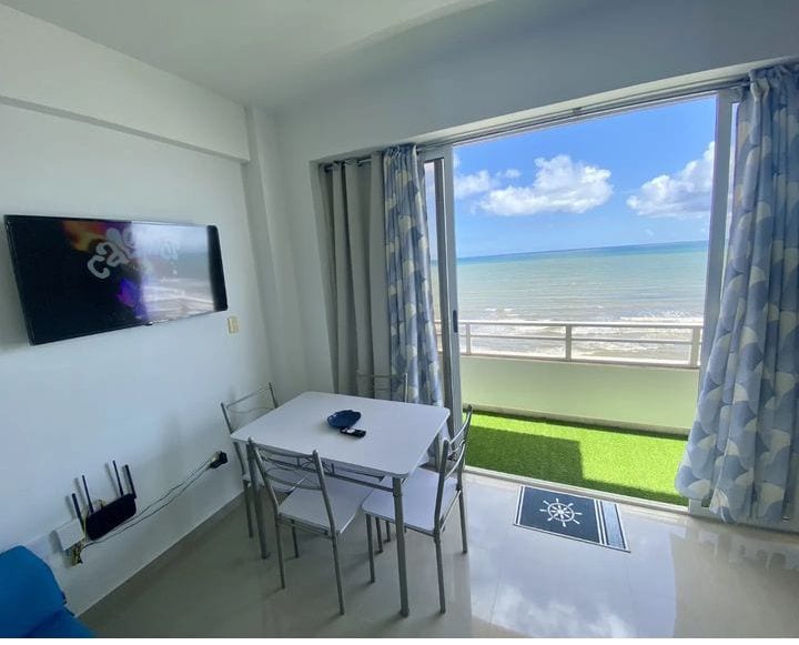 A bright dining area features a white table with four silver chairs, positioned beside large sliding glass doors. The view of the ocean and sky is framed by light blue and white curtains, creating an inviting atmosphere for meals with a coastal backdrop.