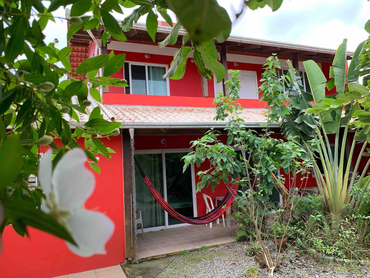 A vibrant red house is framed by lush greenery and tropical plants, with a hammock hung between two supports. The ground level includes a sliding glass door leading to an inviting interior space, while a patio area with chairs is visible in front.