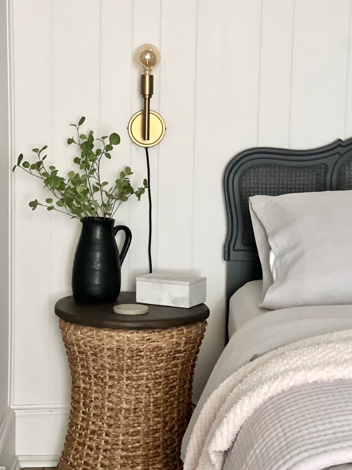 A bedside table made of woven material holds a dark vase with greenery and a small decorative box. A soft gray blanket is draped over a spacious bed, which features a classic headboard and a wall-mounted lamp for ambient lighting.