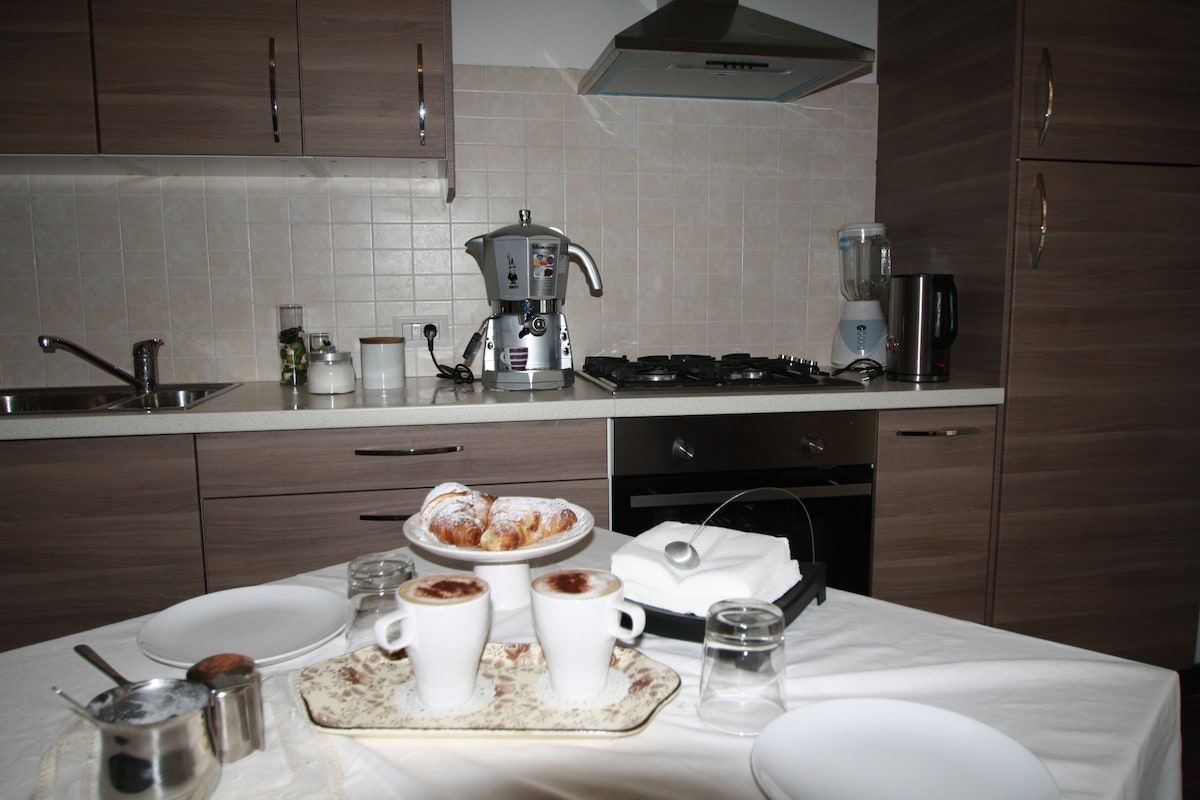 A modern kitchen is visible, featuring sleek cabinetry and essential appliances. A table is set with two cups of coffee and a plate of pastries, inviting a warm dining experience. Countertops are well-organized with a blender and coffee machine, creating a functional cooking space.