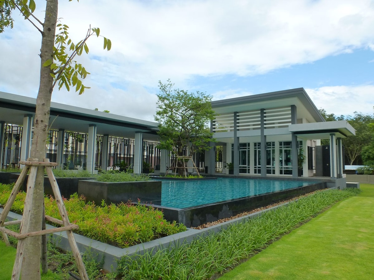 A modern house features large glass windows and a covered entryway. A serene swimming pool is surrounded by landscaped gardens with lush greenery and ornamental plants, providing a tranquil outdoor space. The setting is complemented by a peaceful sky with scattered clouds.