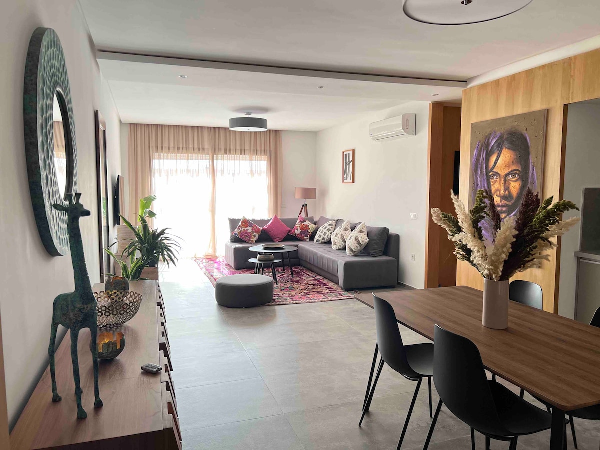 A modern living area is featured, showcasing a spacious gray sofa with colorful cushions. A round coffee table rests in front, surrounded by a cozy seating arrangement. Natural light filters through large windows, creating an inviting atmosphere, complemented by a dining table set for six.