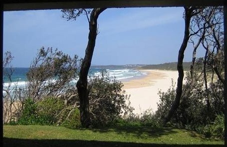 A panoramic view reveals a sandy beach bordered by lush greenery. Waves gently lap at the shore, contrasting against the blue ocean. A clear sky stretches above, with a few scattered clouds, enhancing the serene coastal atmosphere.