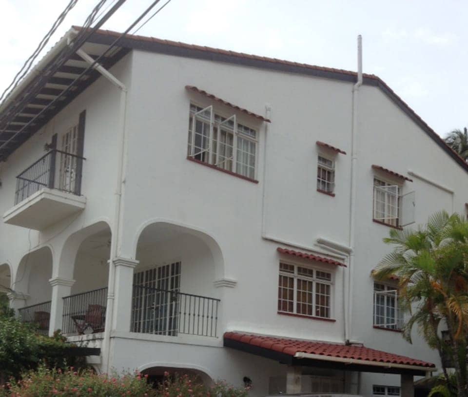 The exterior of a two-story, white townhome showcases a combination of arched and rectangular windows. A red-tiled roof complements the architectural details, while a balcony features black railings. Lush greenery surrounds the home, adding a natural touch to the inviting façade.