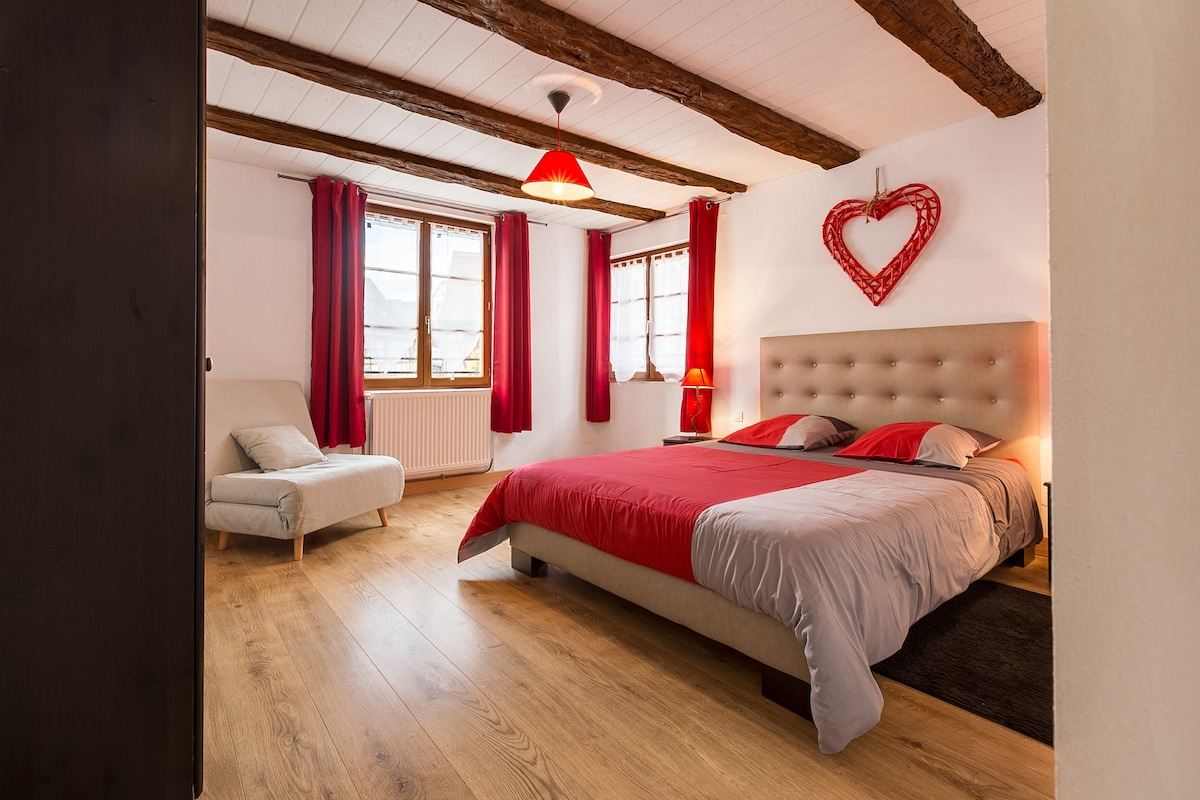 A spacious bedroom showcases a light wood floor and exposed wooden beams on the ceiling. A large bed with a beige headboard is dressed in red and grey linens. Two windows allow natural light, with sheer curtains creating a bright atmosphere. A cozy armchair and decorative heart artwork add charm.
