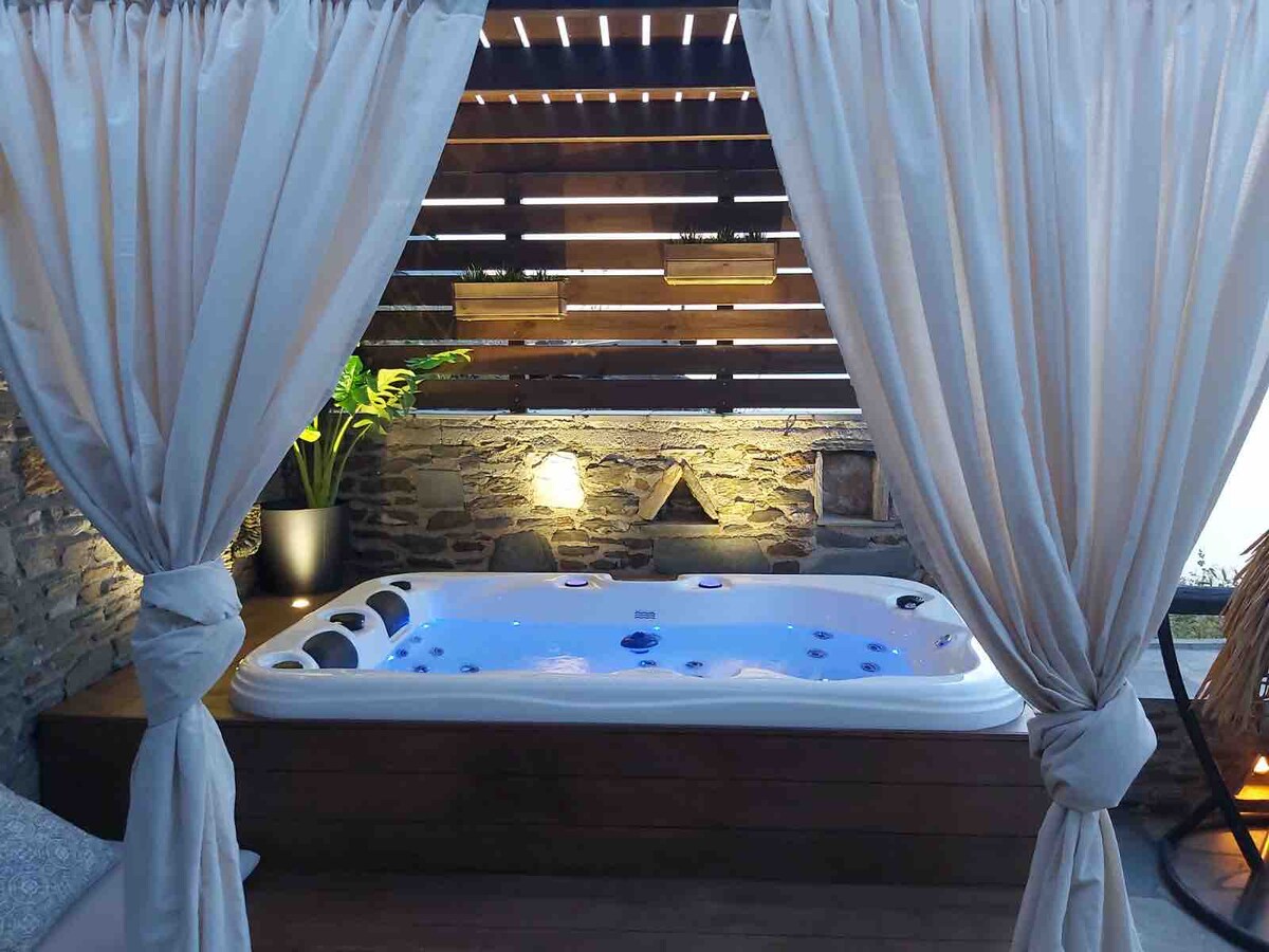 A private jacuzzi is framed by soft, flowing curtains, providing an intimate setting. The jacuzzi is illuminated with a calming blue light, complemented by natural stone walls and wooden accents. Greenery is present in a planter, enhancing the serene ambiance.