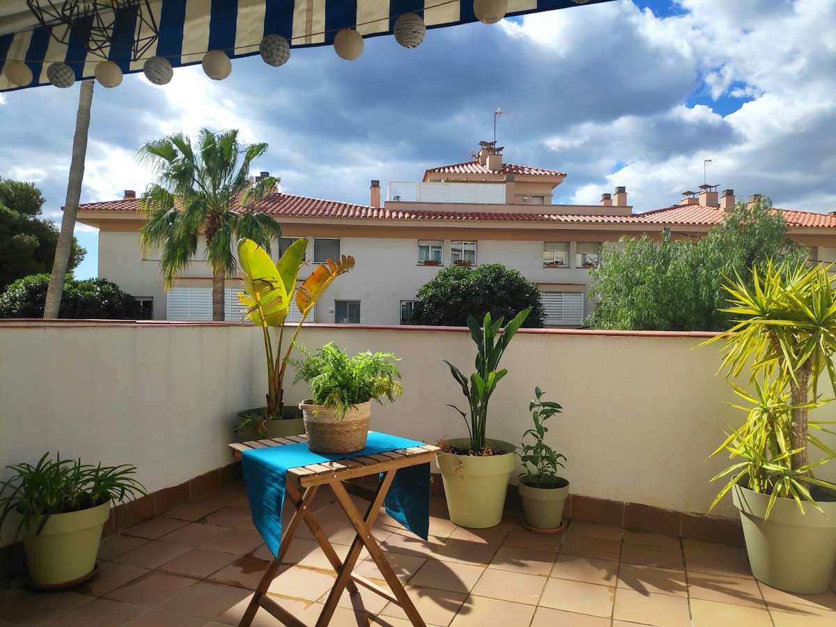 A spacious terrace features a blue table with a potted plant and several leafy green plants in pots around it. A striped awning provides shade, while a view of neighboring buildings and palm trees is visible in the background, creating a calm atmosphere.