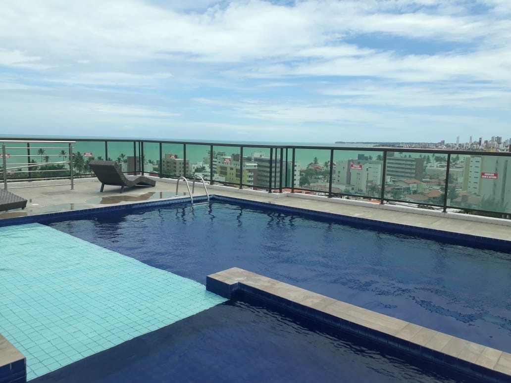 A rooftop pool is showcased with a vibrant blue tile design, offering panoramic views of the sea and nearby cityscape. The deck is bordered by glass railing, ensuring unobstructed sightlines. A single lounge chair rests beside the pool, inviting relaxation in a serene setting.