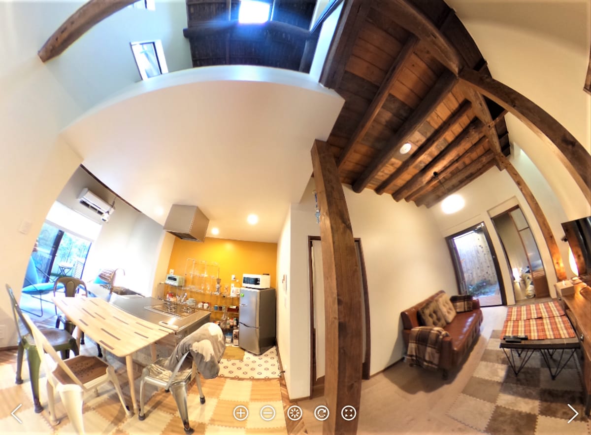 The image captures a spacious interior featuring an open dining area with a wooden table surrounded by metal chairs. Natural light fills the space, illuminating the warm orange wall across from a modern kitchen. Exposed wooden beams add character overhead, while a cozy seating area is visible in the background.